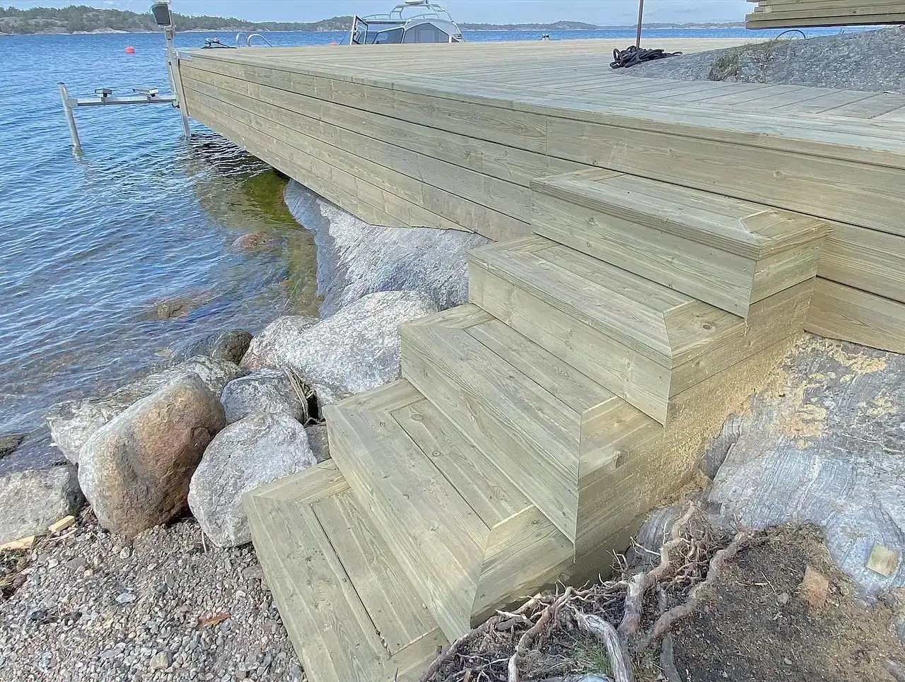 Trätrappa leder ner från brygga till stenig strand vid vatten.