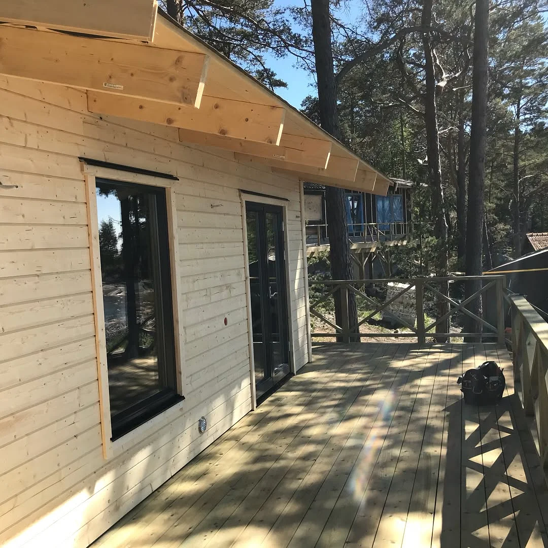 Träaltan med glasdörrar vid trähus omgivet av tallskog.