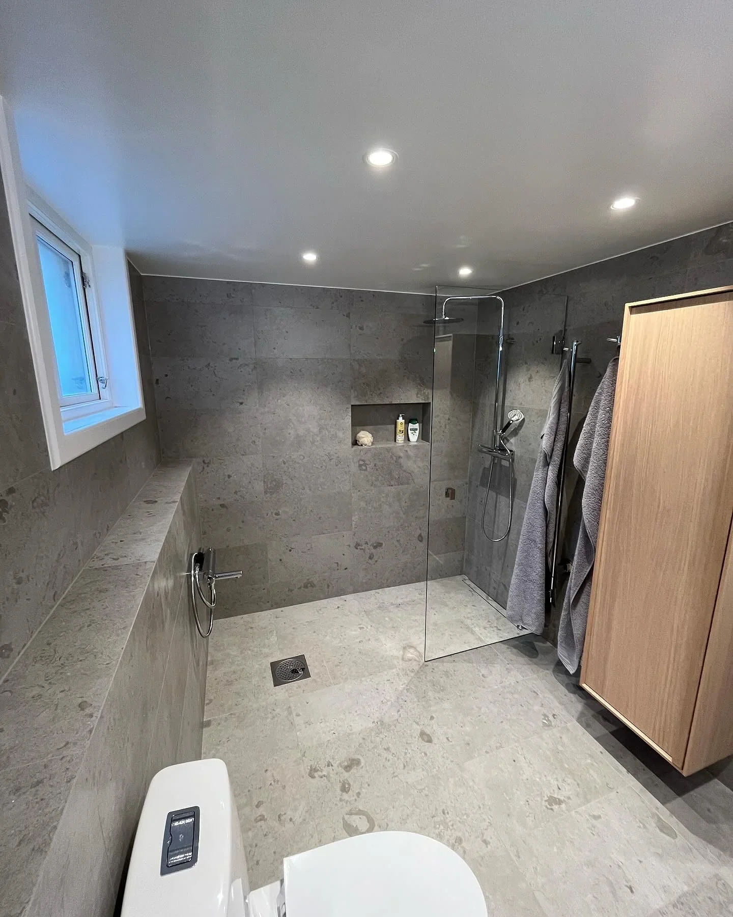 Modernt badrum med grå stenplattor, glasduschwand och träskåp.