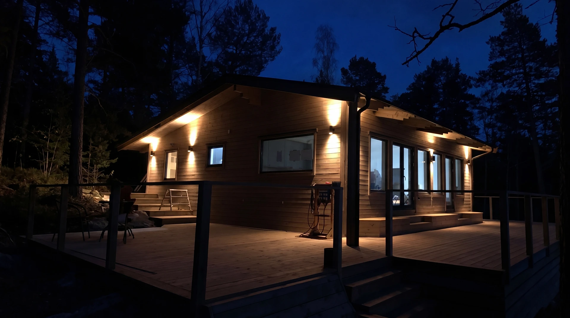 Belyst trähus med stora fönster i skogsmiljö vid skymning.