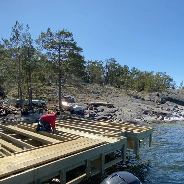 Person bygger brygga vid klippstrand med trävirke under solig dag.