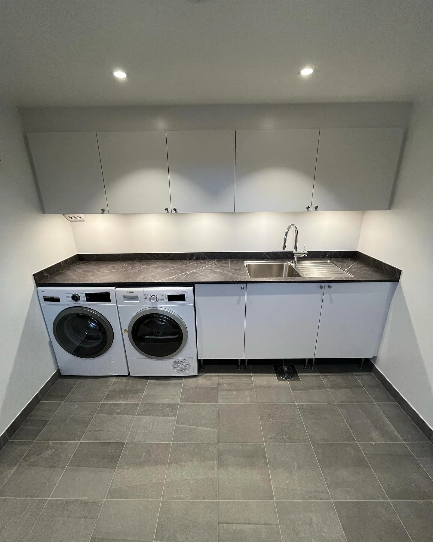 Modern tvättstuga med vit inredning, tvättmaskin, torktumlare och diskbänk.